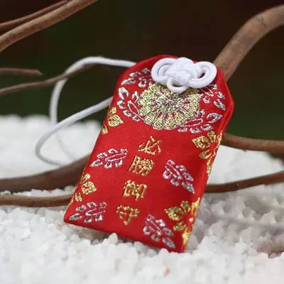 Breloque omamori en soie rouge avec des motifs décoratifs dorés et argentés et un nœud blanc.